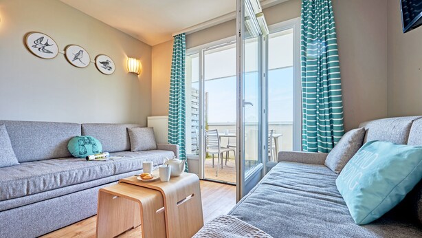 A modern living room with a grey sofa, a wooden side table, and a balcony with striped curtains. - Residence Pierre & Vacances Le Chant des Oiseaux (Courseulles-sur-Mer)