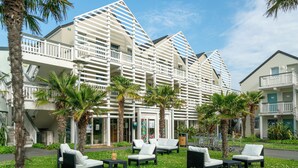 A hotel with a modern architectural design, outdoor seating area, and palm trees. - Residence Pierre & Vacances Le Chant des Oiseaux (Courseulles-sur-Mer)