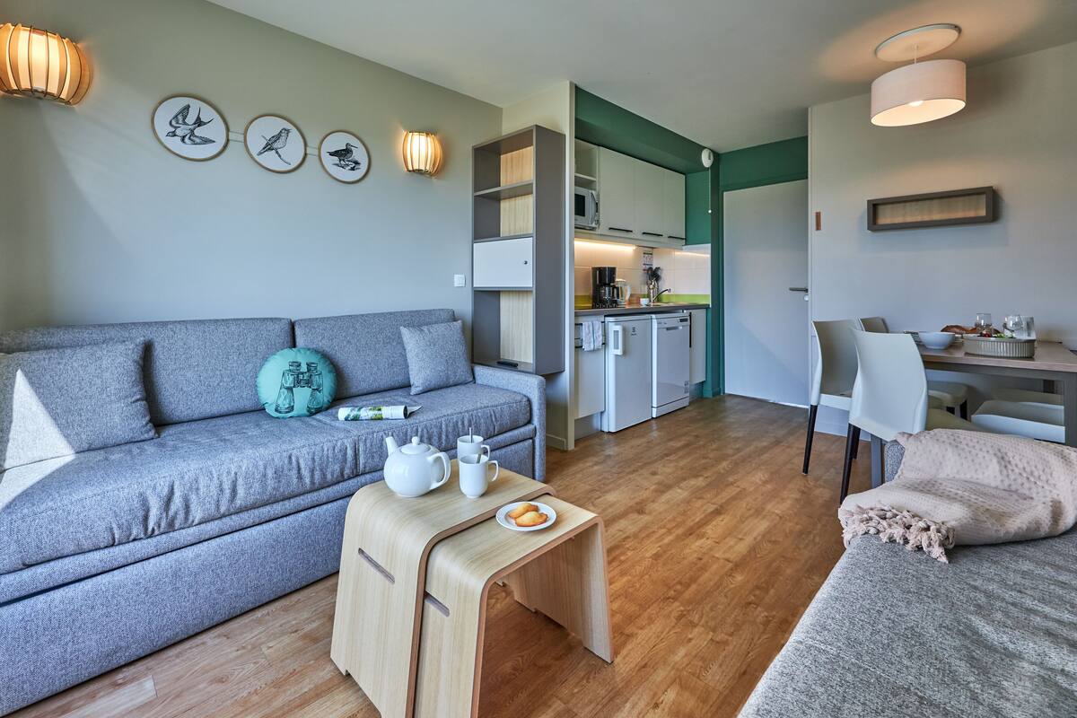 A modern living room with a grey sofa, a small wooden table with a teapot and cup, a dining table with chairs, and a kitchen area with cabinets and a microwave.
