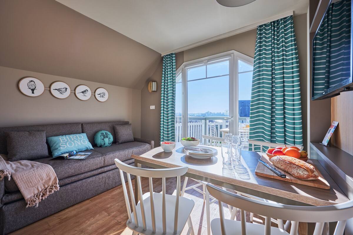 A cozy living area with a sofa, a dining table with chairs, a window with curtains, and wall art.