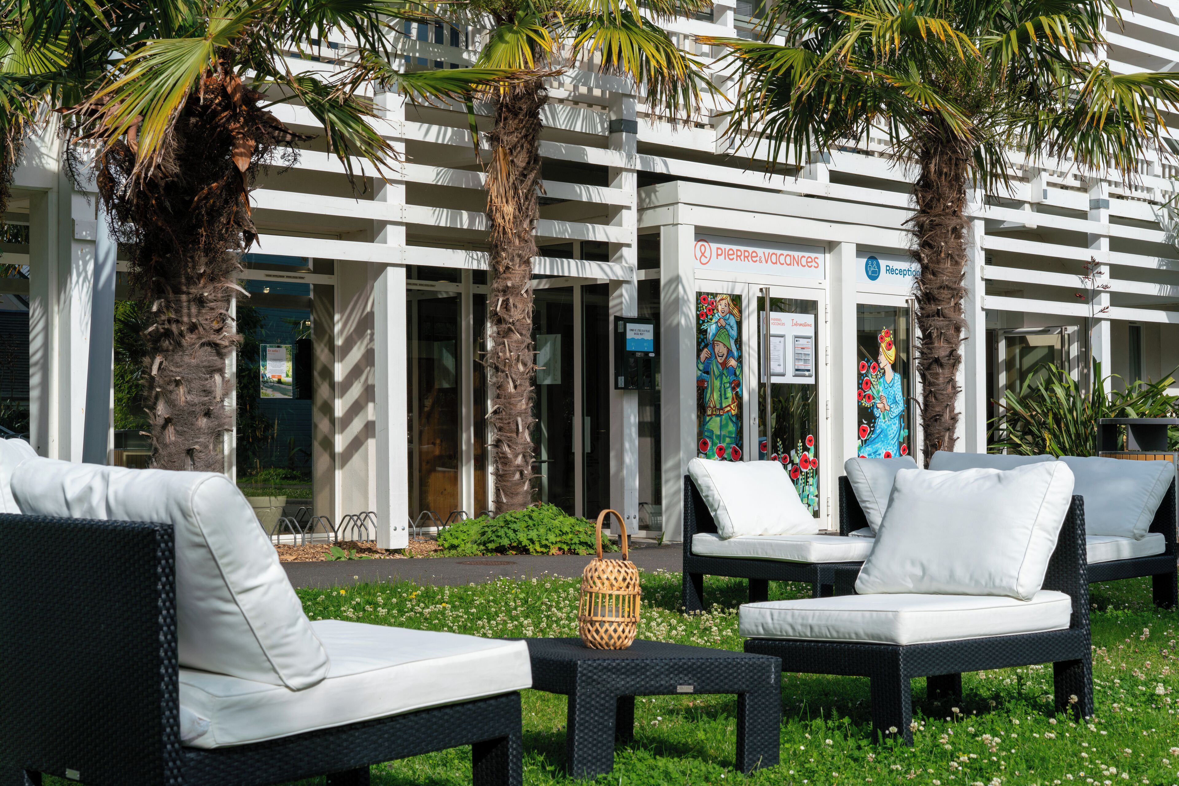 Outdoor seating with white cushions, a wicker table, and palm trees in front of a building with a reception area.