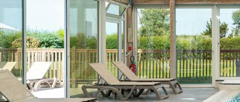 A covered outdoor pool area with lounge chairs, glass doors, and a view of the surrounding greenery.