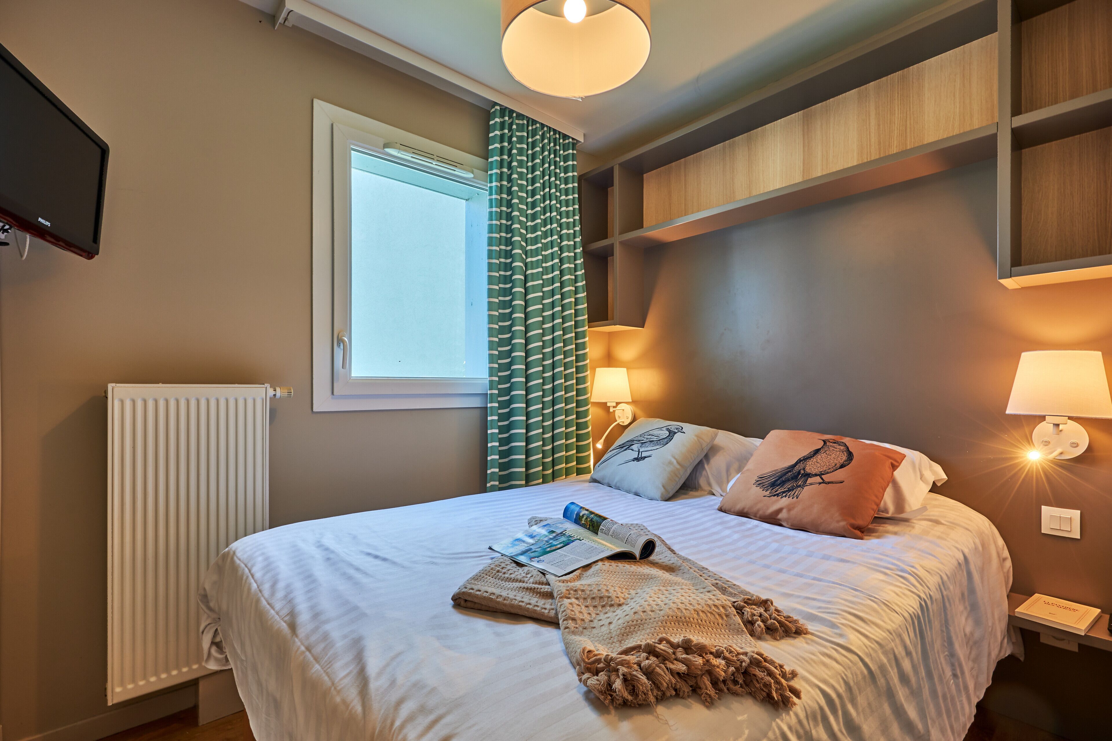 A neatly made bed with a magazine, a knitted blanket, and pillows, including one with a bird design. There is a window with curtains, a wall-mounted TV, and a shelf with a lamp.