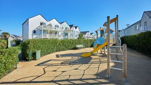 A playground with a slide and climbing structure, surrounded by a pathway and residential buildings. - Residence Pierre & Vacances Le Chant des Oiseaux (Courseulles-sur-Mer)