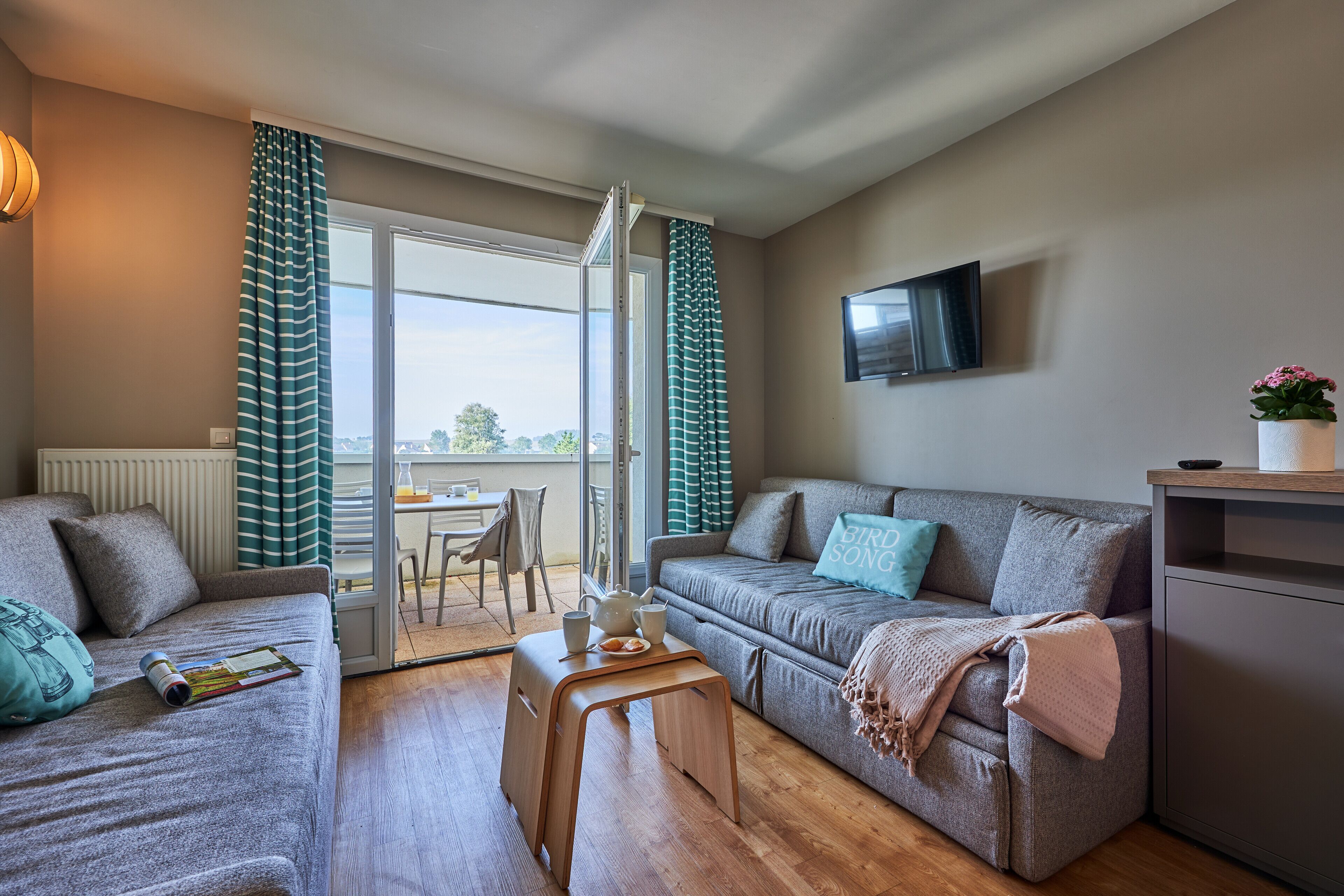 A modern living room with a grey sofa, a wooden coffee table, and a balcony with a view.