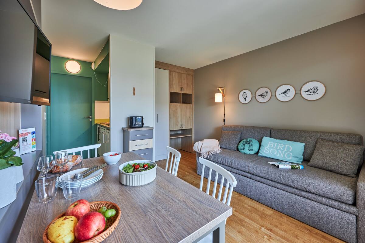 A modern living room with a grey sofa, a dining table with fruits and a bowl of salad, and a kitchen area with wooden cabinets.
