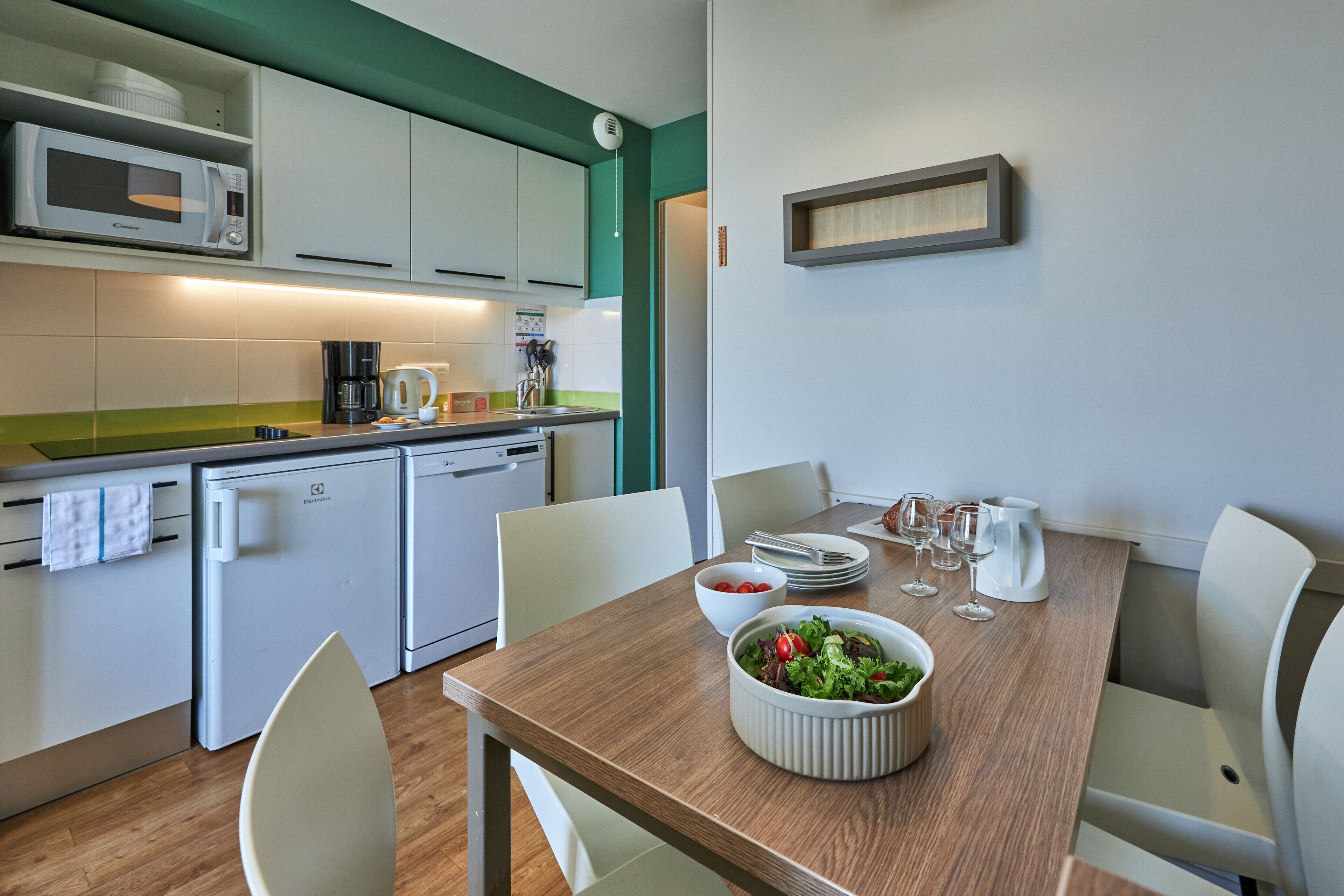 A modern kitchen with white cabinets, a microwave, a coffee maker, and a dining table set with plates, glasses, and a bowl of salad.