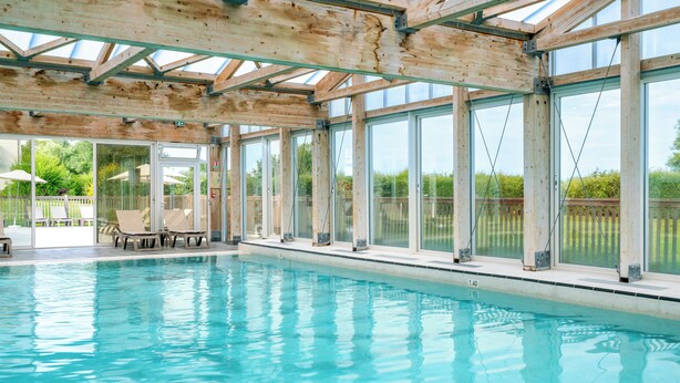 An indoor swimming pool with clear glass walls, wooden beams, and lounge chairs. - Residence Pierre & Vacances Le Chant des Oiseaux (Courseulles-sur-Mer)