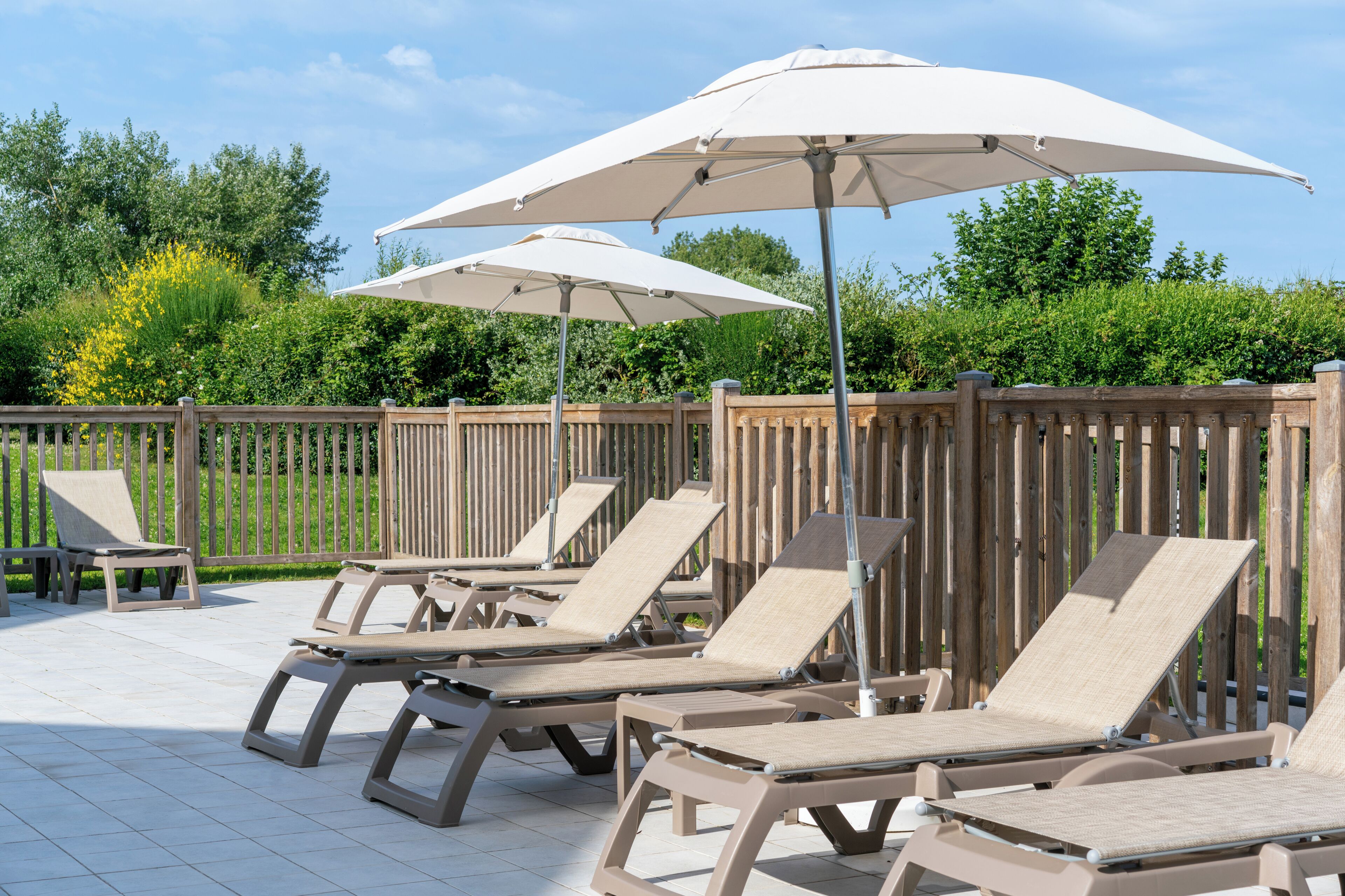 A patio with sun loungers and umbrellas, wooden fence, and greenery in the background.