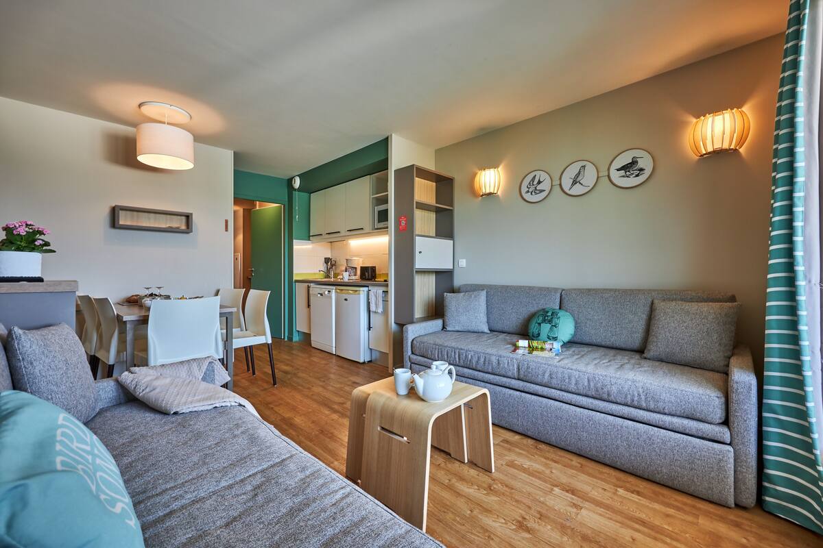 A modern living room with a grey sofa, a wooden coffee table, and a dining area with white chairs.
