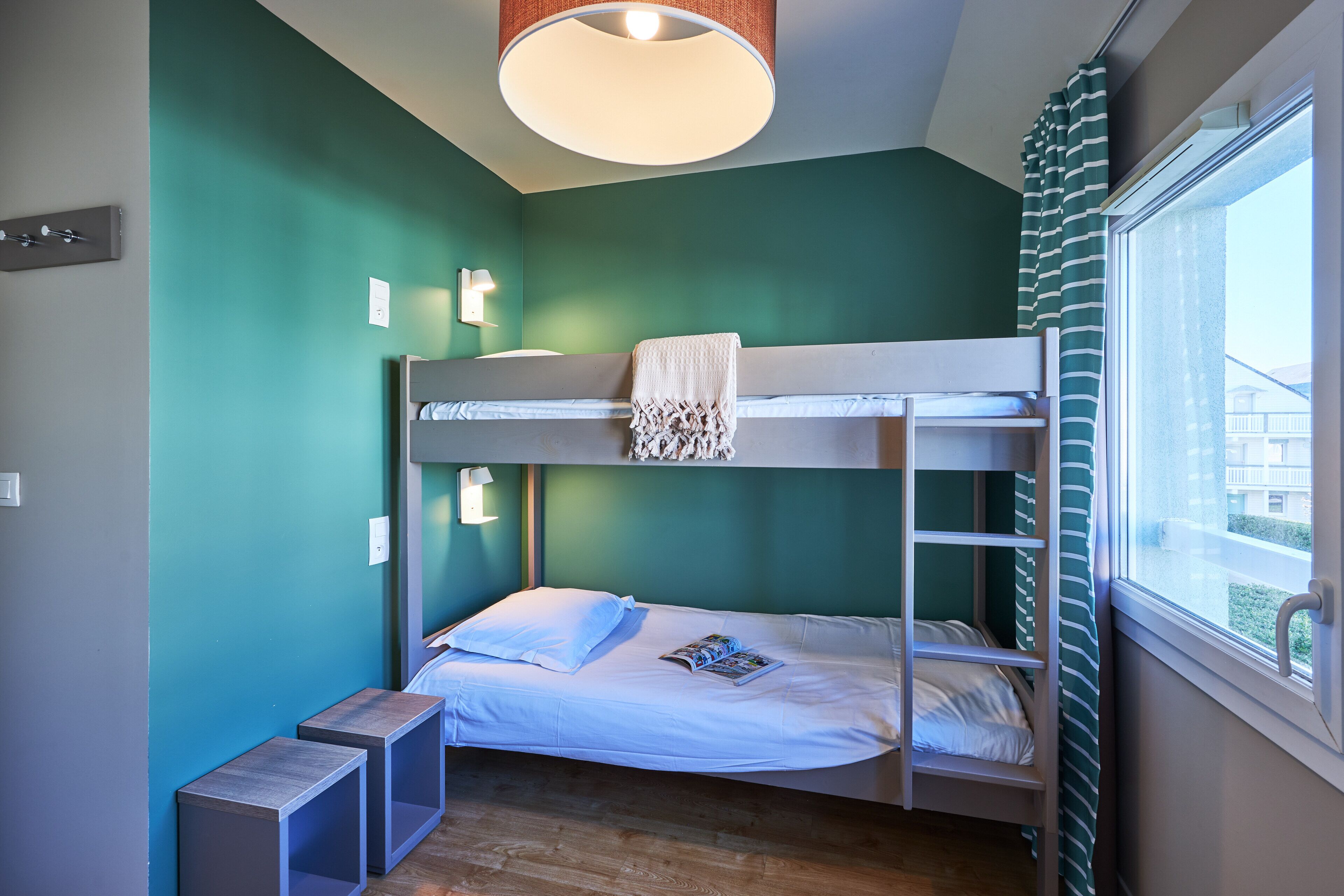 A bunk bed room with a window, a bedside table, and a lamp.