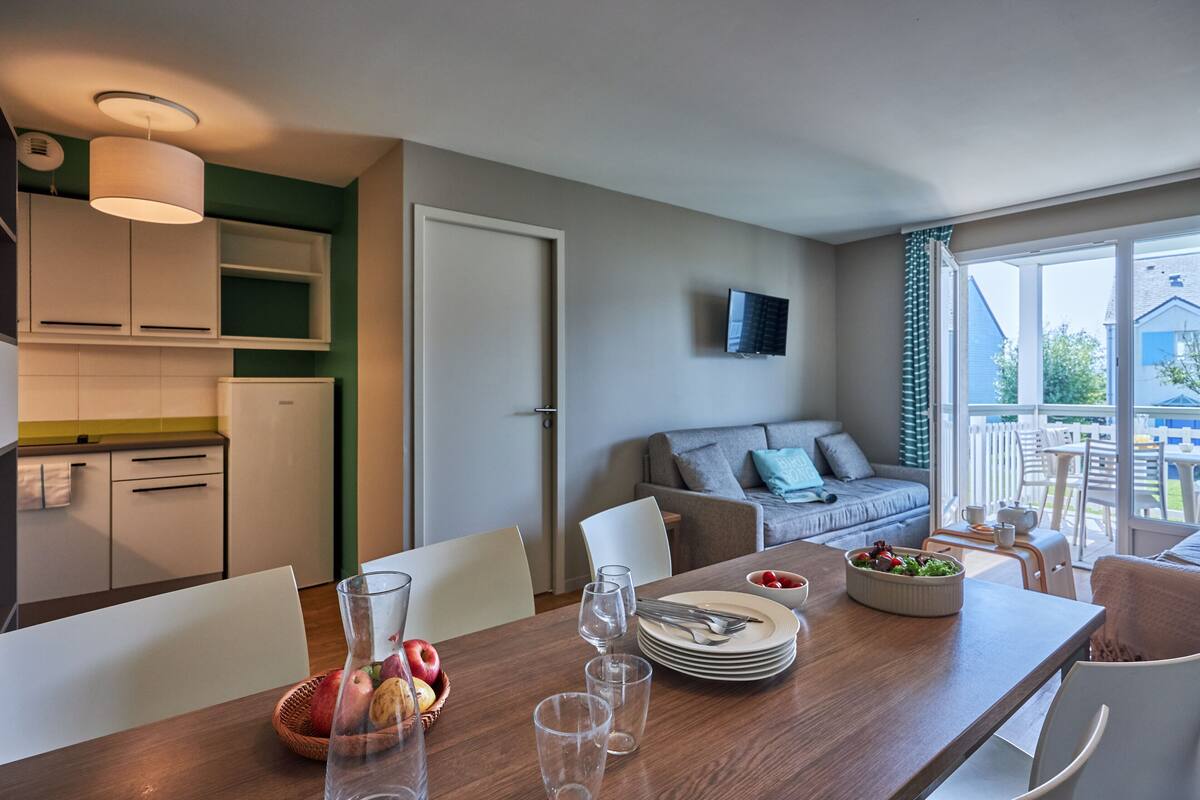A modern kitchen with white cabinets, a dining table set with plates, glasses, and a fruit bowl. There is a sofa and a TV mounted on the wall.