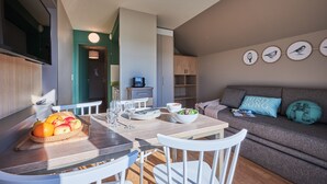 A modern living room with a sofa, a dining table with chairs, and a kitchen area with a TV and cabinets. - Residence Pierre & Vacances Le Chant des Oiseaux (Courseulles-sur-Mer)