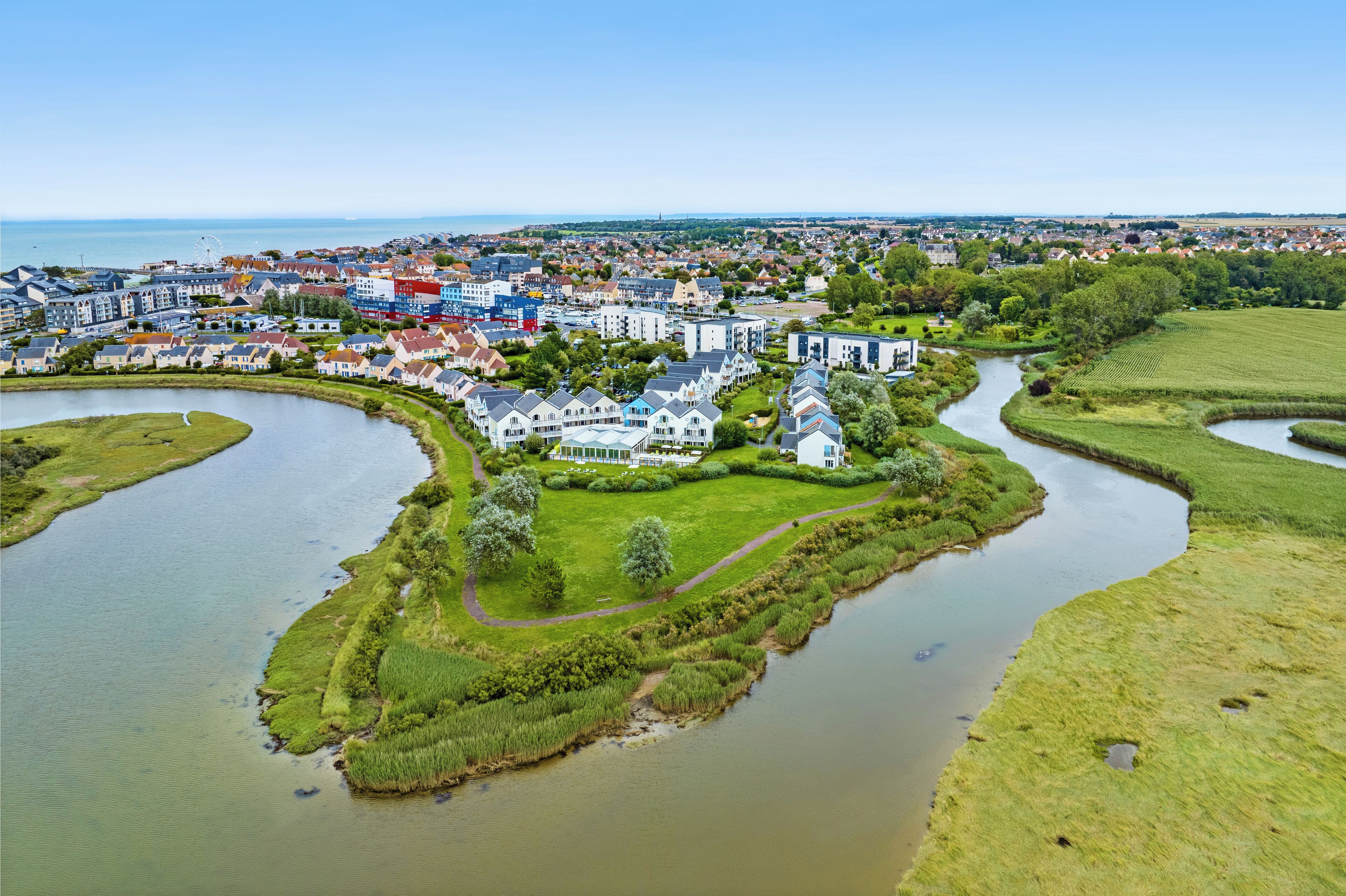 Une vue aérienne d’une ville côtière, avec un imposant bâtiment blanc entouré de champs verdoyants et traversée par une rivière.