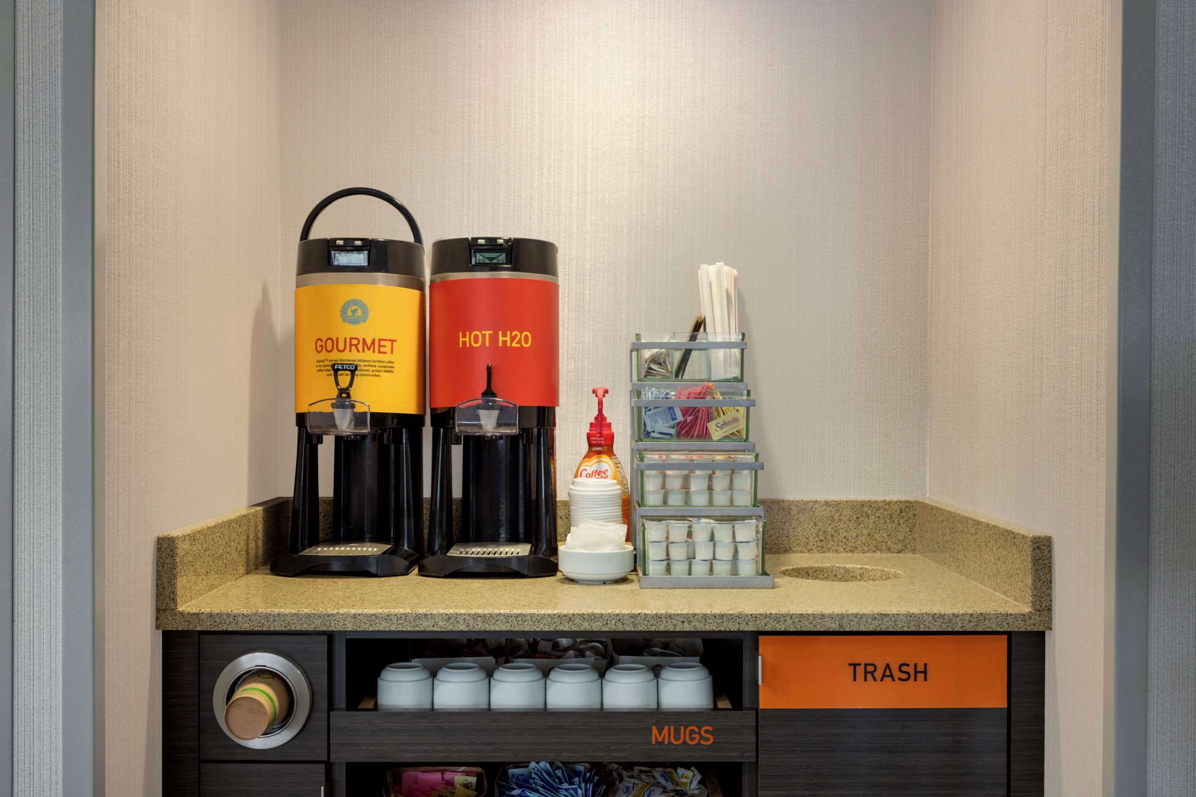 Eine Kaffeestation mit zwei Kaffeeautomaten, einem Gewürzspender und einem Serviettenhalter. Darunter befinden sich beschriftete Schränke für Müll und Tassen.