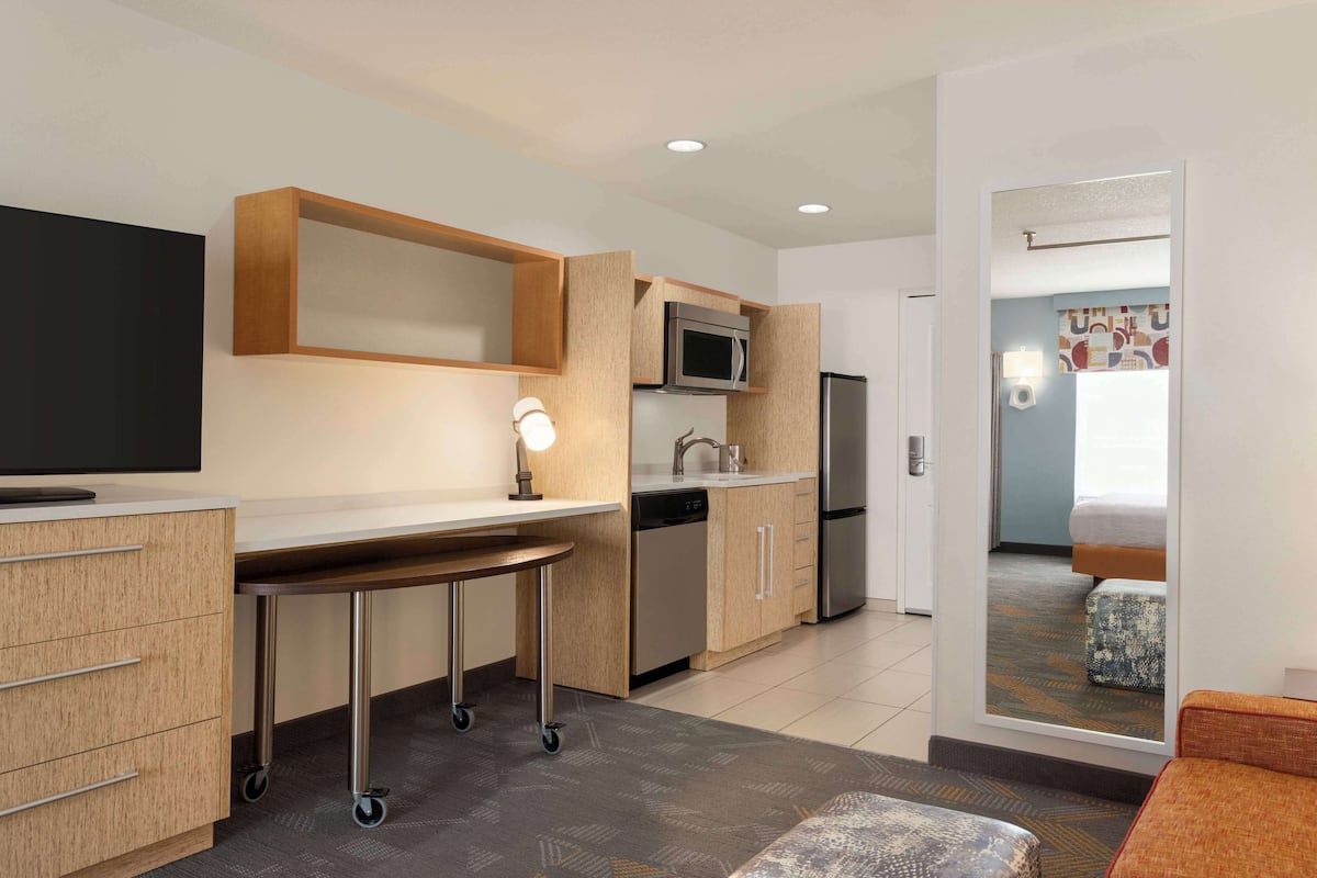A compact hotel room with a kitchenette, a flat-screen TV, a small table, and a sofa.