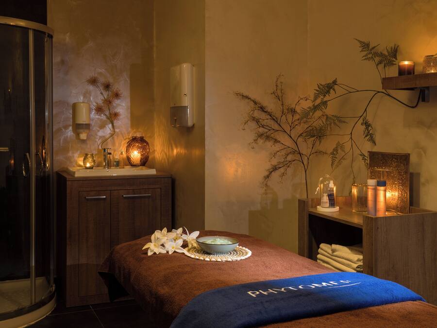 A spa room with a massage table, a glass shower enclosure, a wooden cabinet, and a shelf with candles and a vase.