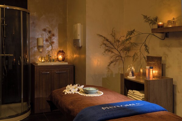 Un cuarto de spa con una camilla de masaje, cabina de ducha de vidrio, un armario de madera y un estante con velas y un florero.
