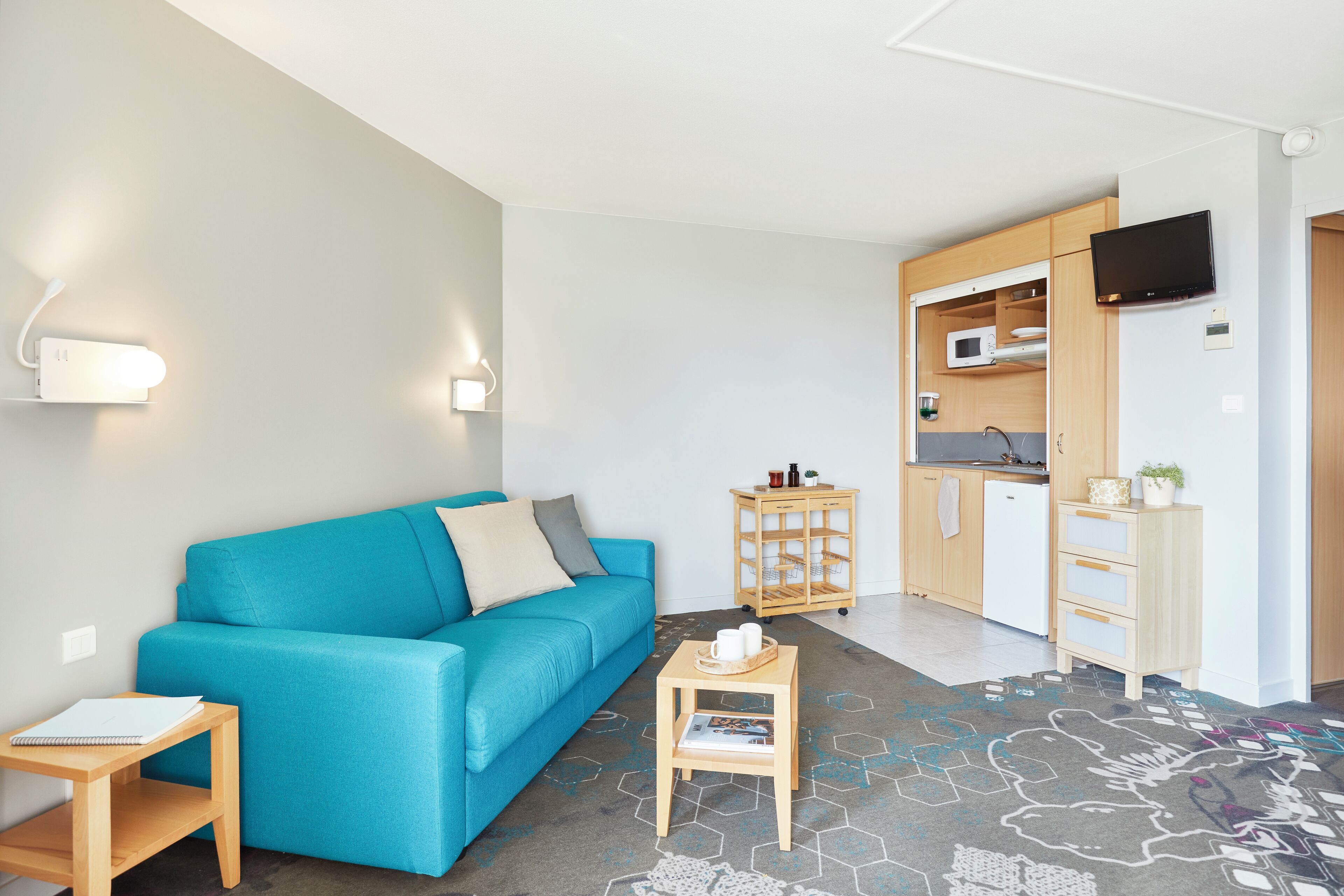 Un salon moderne avec un canapé de couleur turquoise, une petite table d’appoint en bois et un coin cuisine équipé d’un four à micro-ondes et de placards.