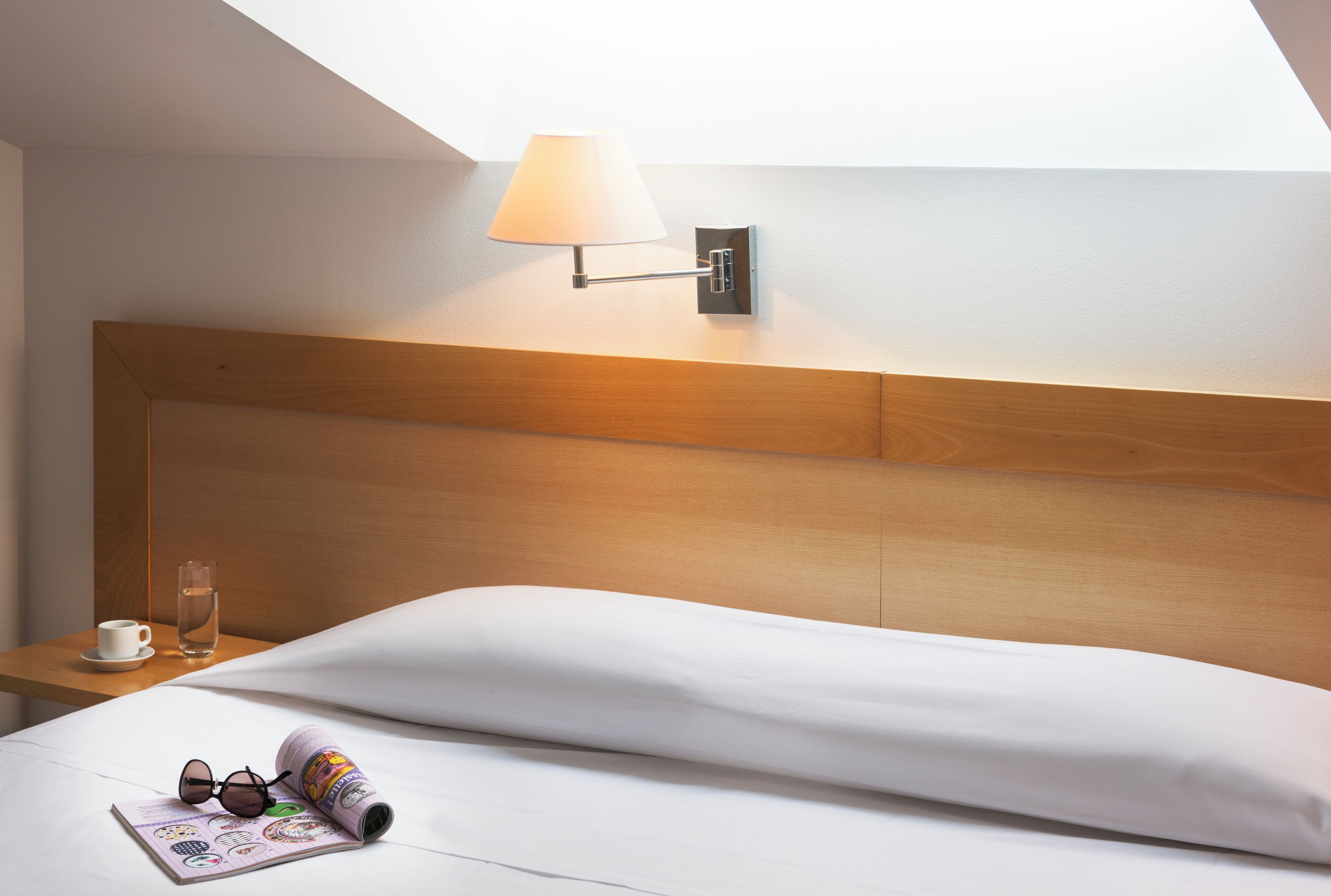 Hotelkamer met een houten hoofdeinde, een nachtkastje met een beker en een glas water, een opgerold tijdschrift en een paar zonnebrillen.