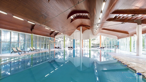 A large indoor swimming pool with wooden ceiling beams, clear glass walls, and lounge chairs along the poolside.
