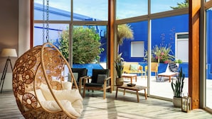 A spacious room with a hanging chair, wooden furniture, and large windows overlooking a blue building and greenery.