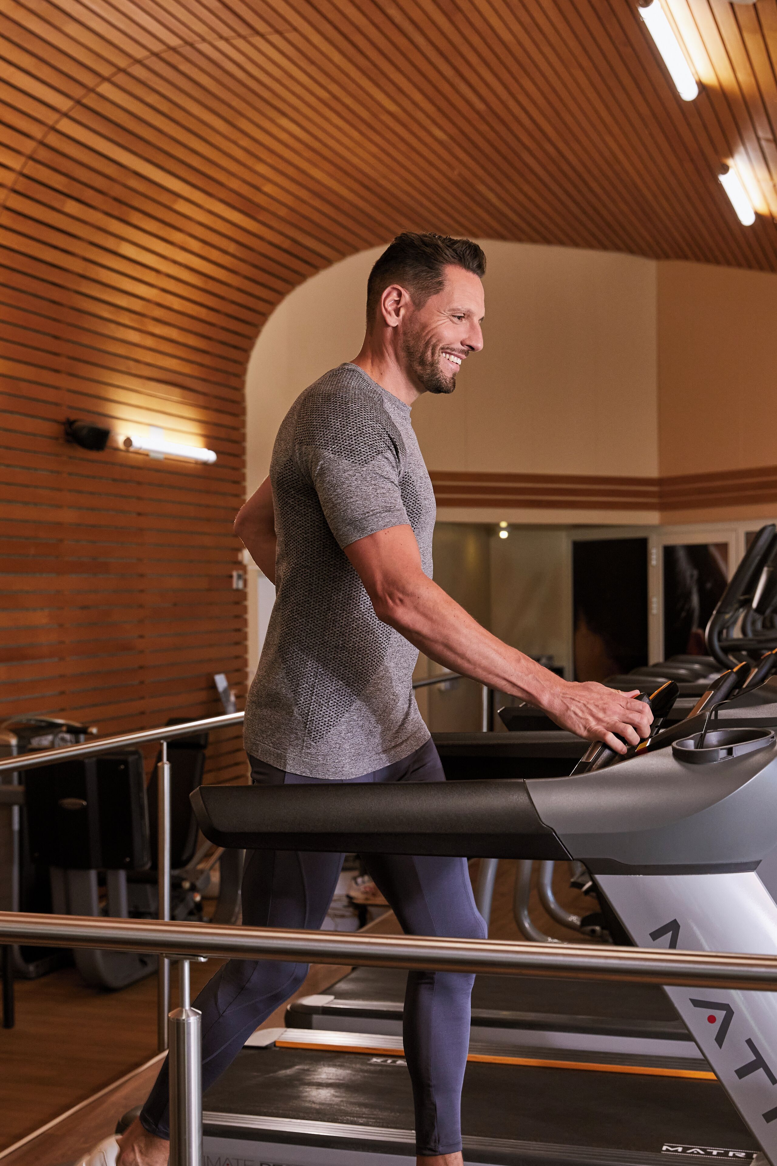 Une personne utilise un tapis de course dans une salle de sport. Le plafond est en bois et on aperçoit, au fond, du matériel d'exercice.
