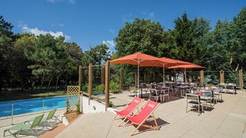 Un espace aménagé au bord de la piscine, avec des chaises longues et des parasols, entouré d’arbres et donnant sur la piscine.
