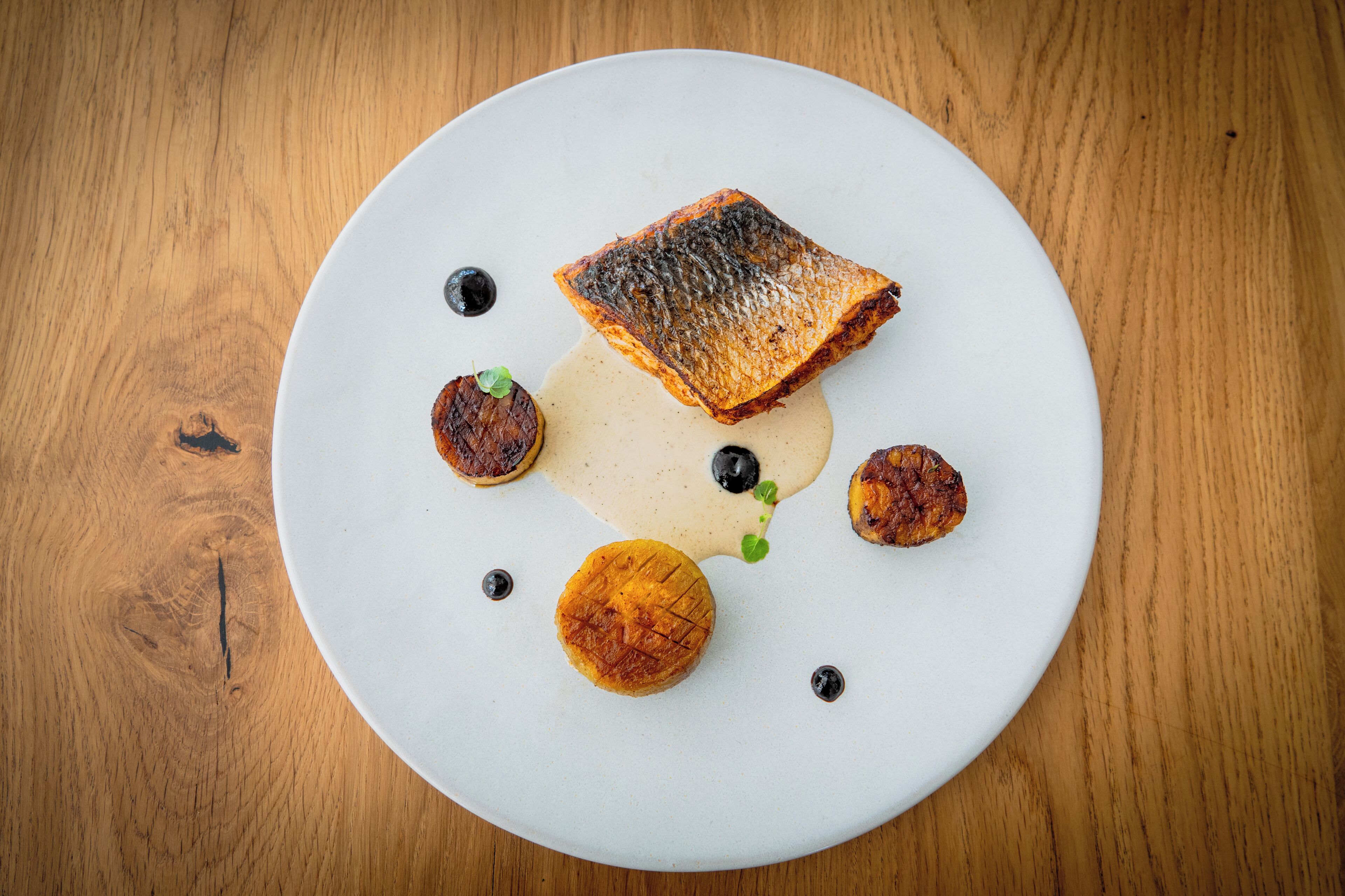 Un plato blanco con un pescado a la parrilla, una porción de salsa y varios acompañamientos redondos sobre una mesa de madera.