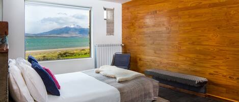 A bedroom with a large window, a bed with pillows, a chair, a radiator, and a wooden wall.