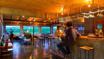 Un restaurante con interior de madera, zona de bar y amplios ventanales con vistas a un paisaje verde.