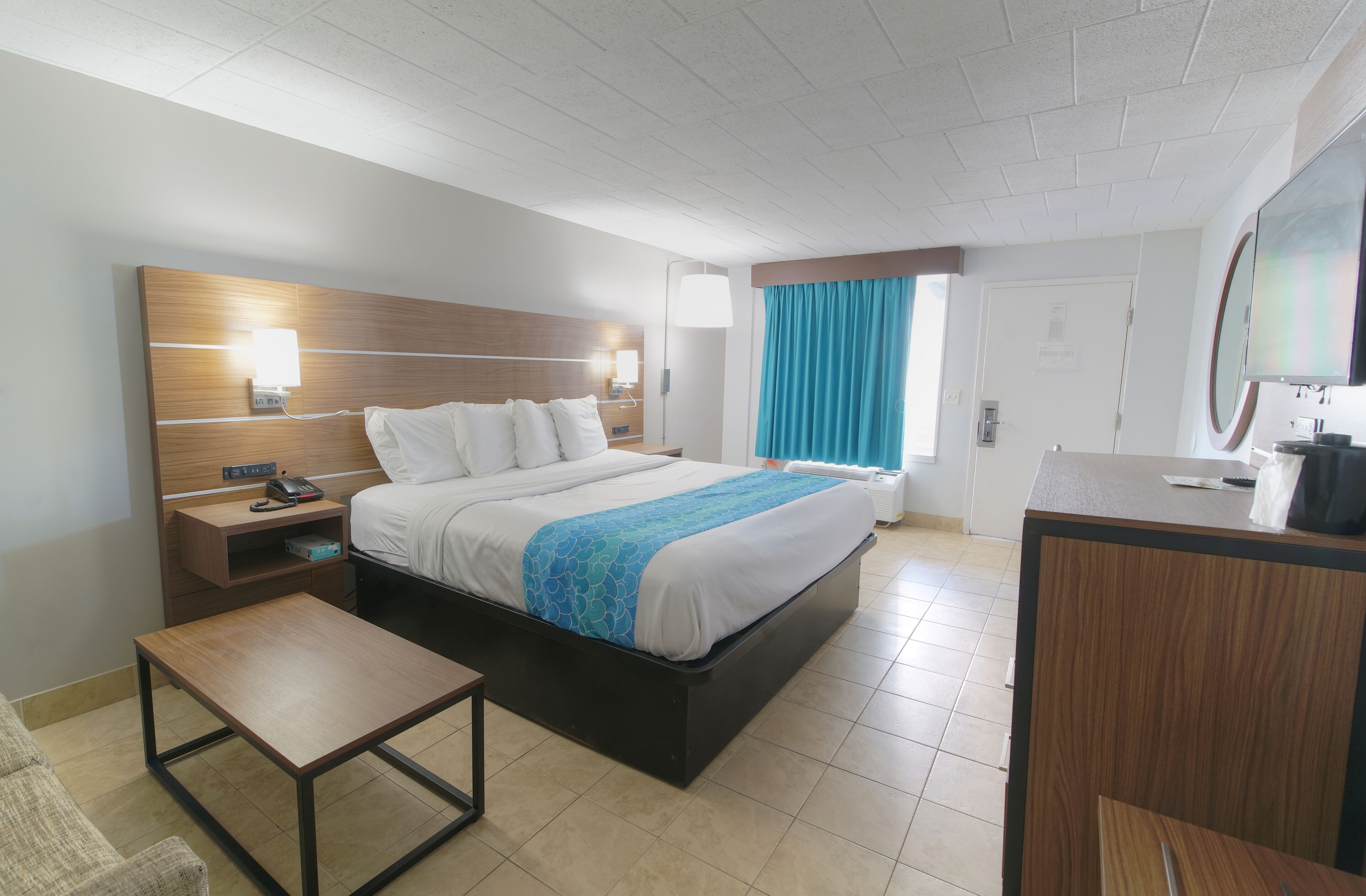 A hotel room with a bed, bedside table, lamp, and a desk with a chair.