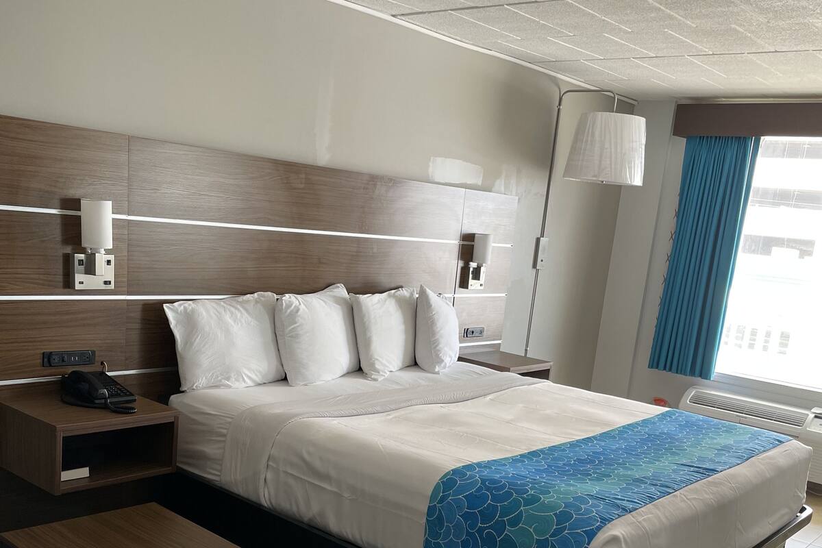 A hotel room with a bed, bedside table, lamp, and a view of the outside.