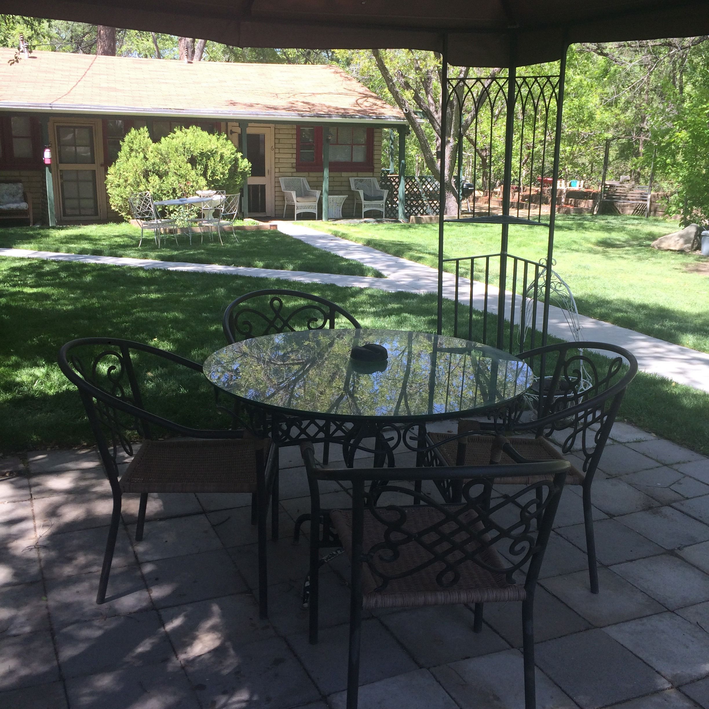 A patio with a round table and chairs, surrounded by a garden with a lawn and trees.
