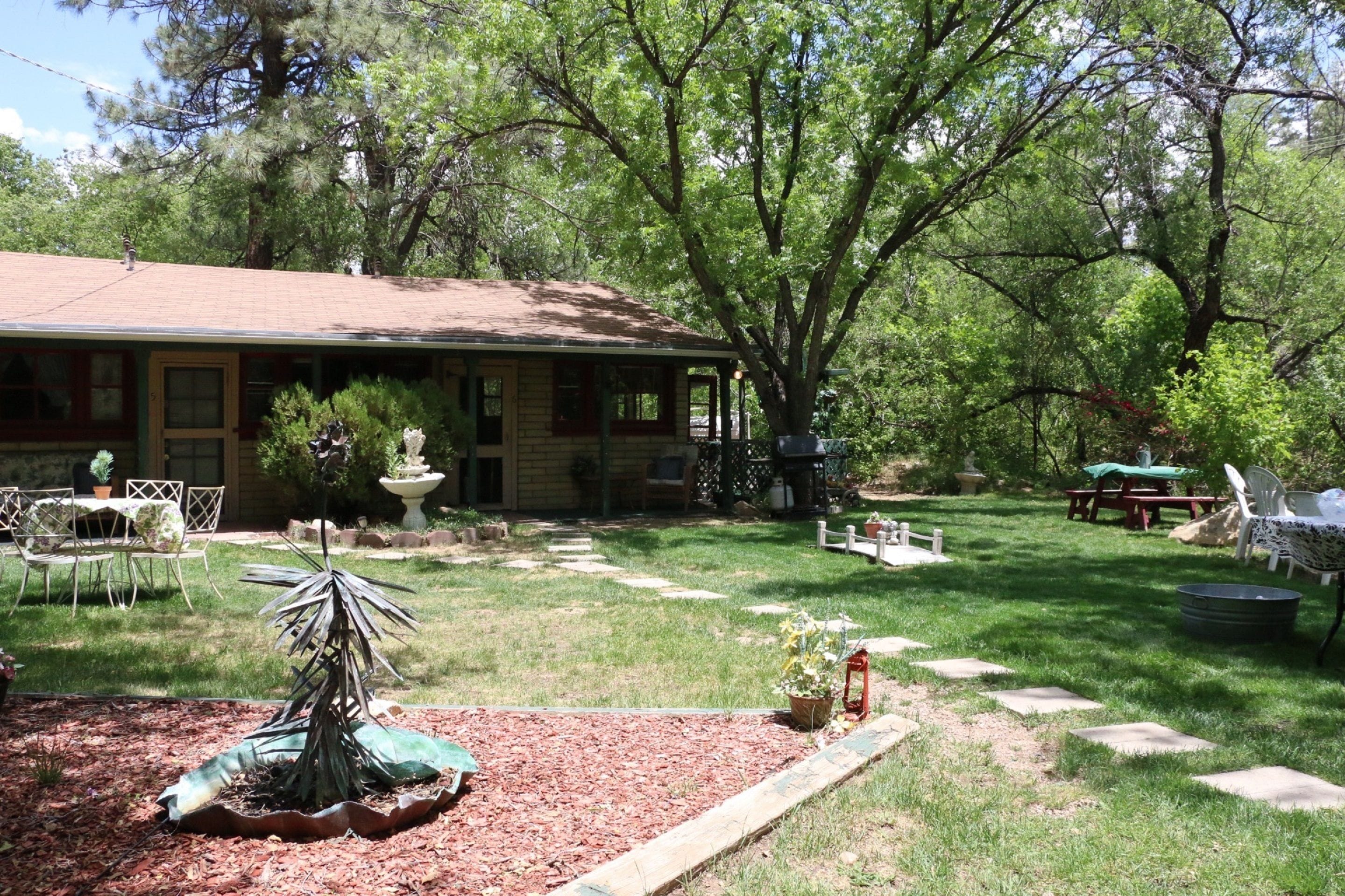 Una casa con un porche cubierto, un jardín con un árbol pequeño y una fuente, y una mesa de picnic en el patio.