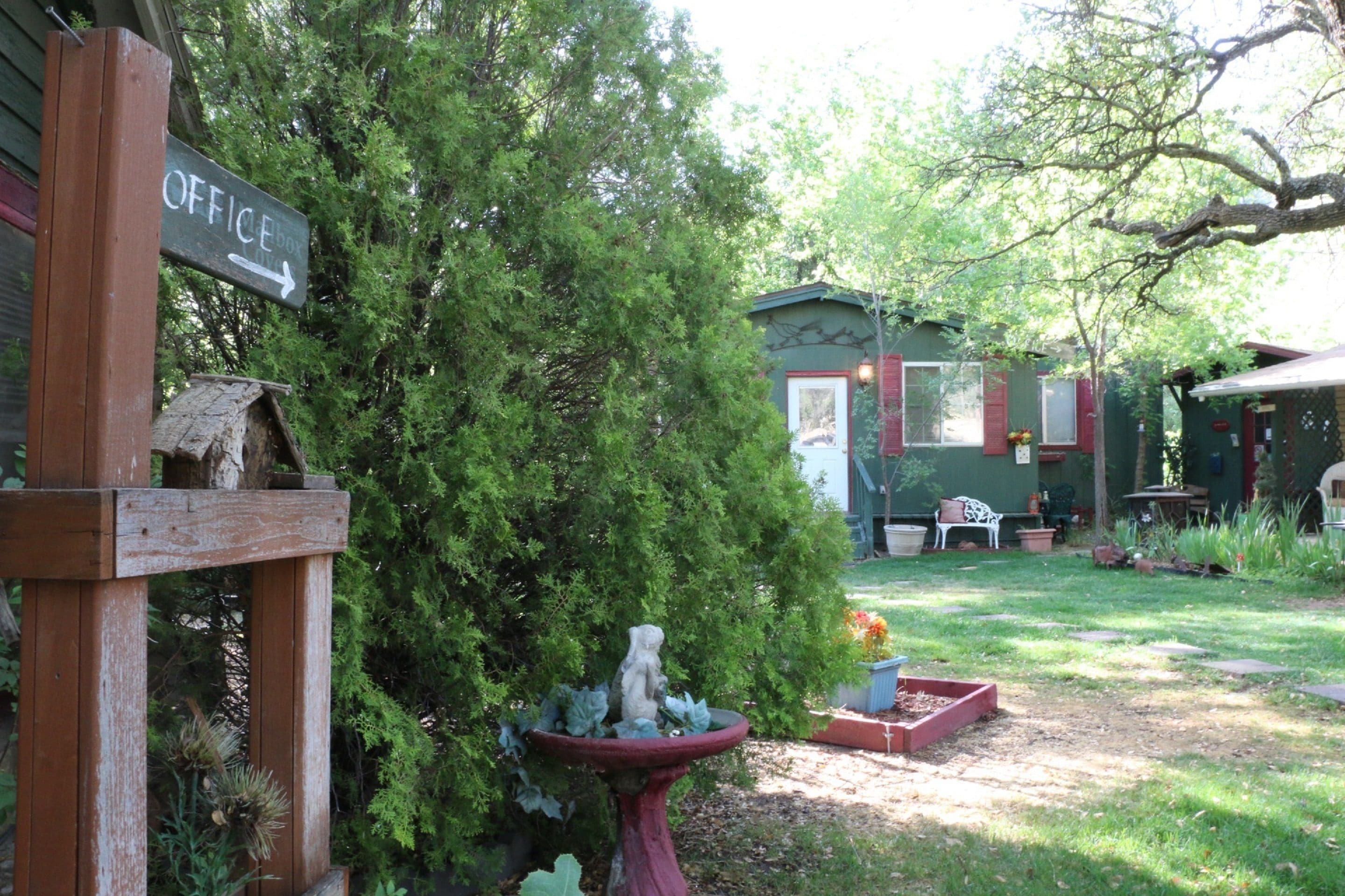 Un panneau en bois indiquant « BUREAU » avec une flèche pointant vers la droite, une nichoir en bois, un abreuvoir pour oiseaux et un petit abri de jardin, le tout dans un jardin arboré avec un banc en arrière-plan.