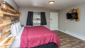 A bedroom with a wooden headboard, a large bed with a pink blanket, a TV mounted on the wall, and a window with curtains. - Apple Creek Cottages (Prescott)