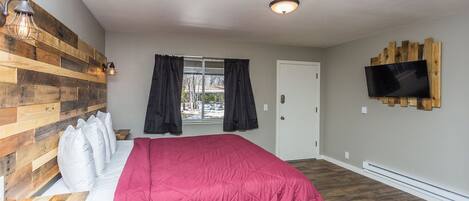 A bedroom with a wooden headboard, a large bed with a pink blanket, a TV mounted on the wall, and a window with curtains.