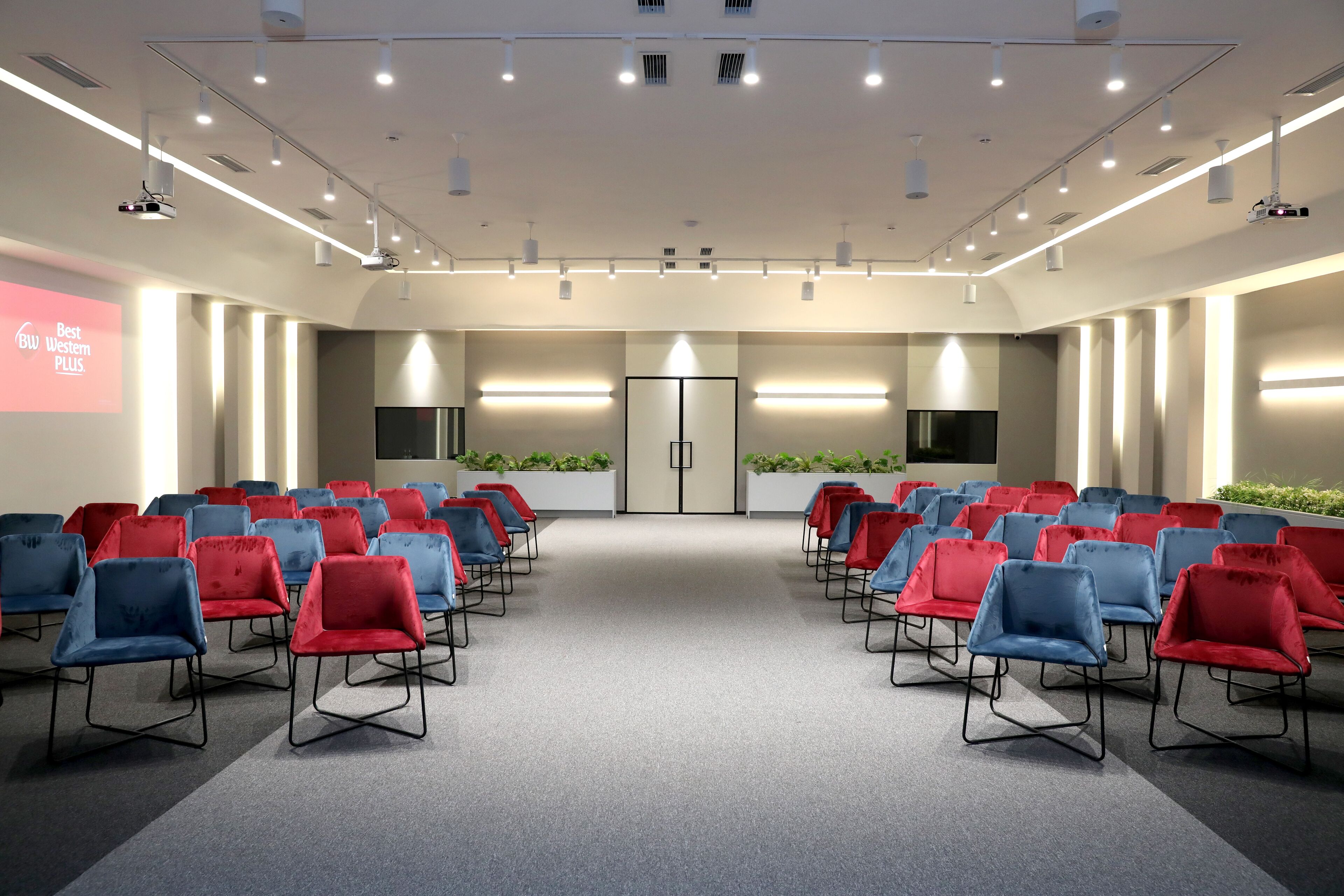 A conference room with rows of red and blue chairs, a projector screen, and a large door at the front.