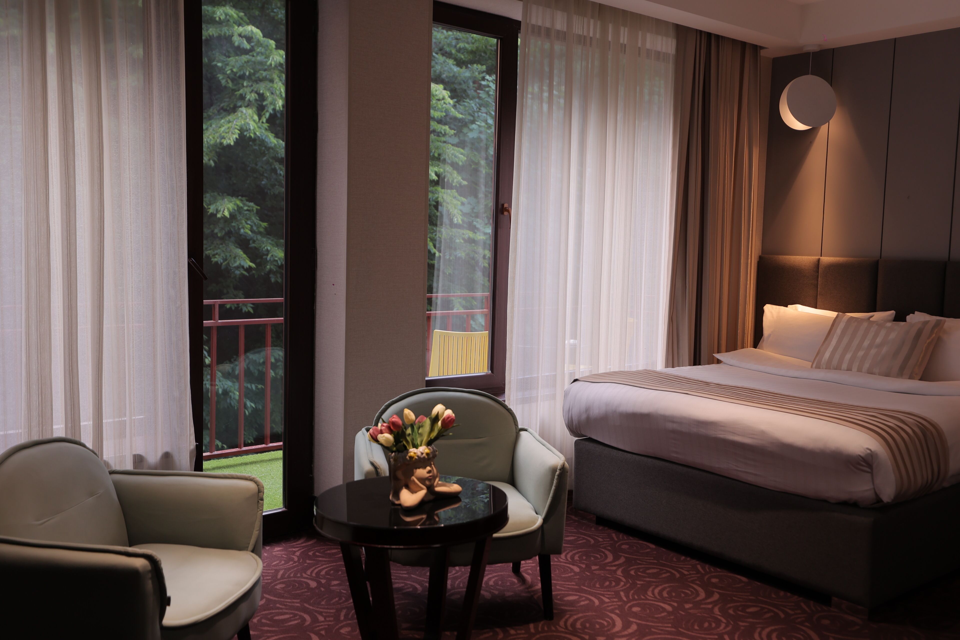 Een hotelkamer met een bed, twee stoelen, een tafel met een vaas, en uitzicht op bomen door het raam.