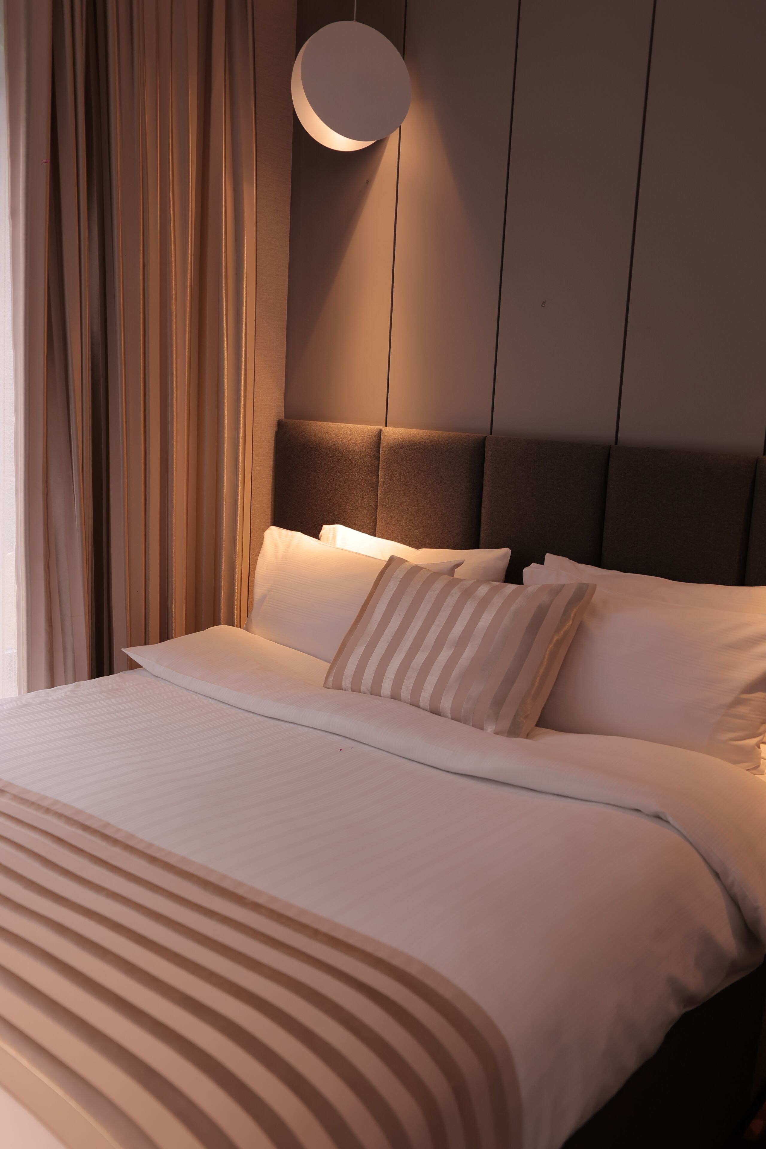 Una cama bien hecha con un cojín a rayas, una lámpara de pared redonda y un cabecero.