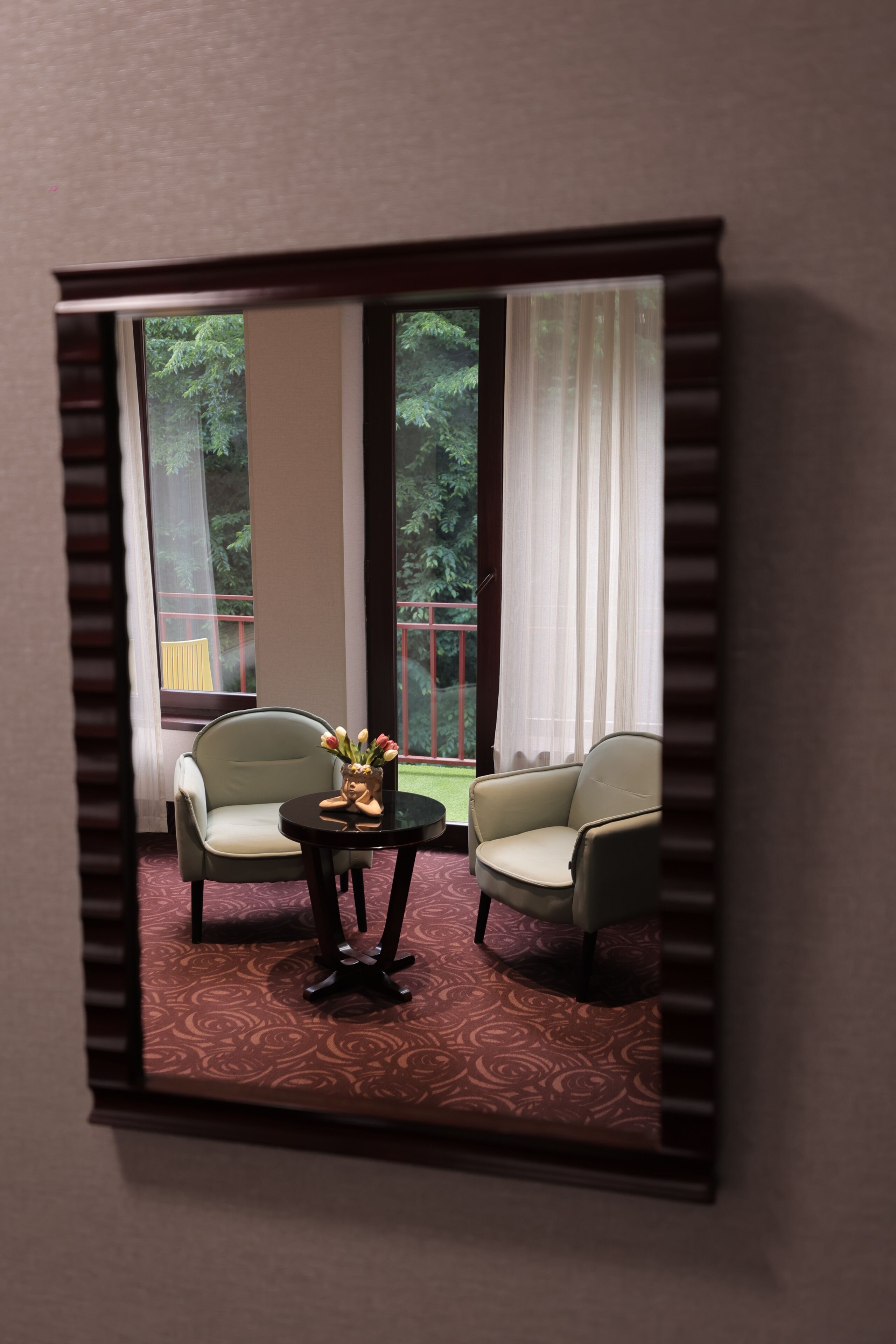 A room with two armchairs, a small table with a vase, and a view of a balcony with greenery.
