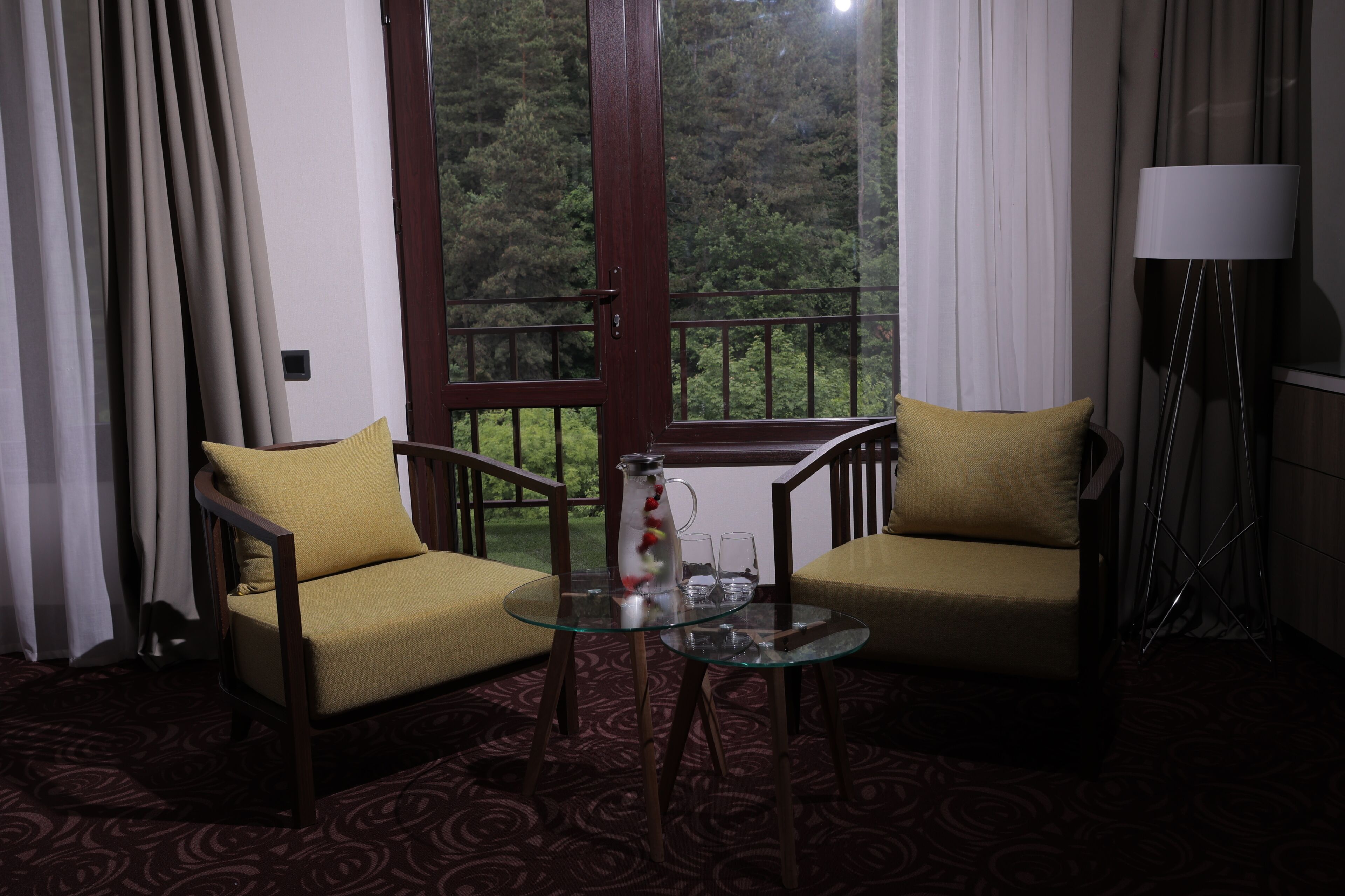 A room with two armchairs, a glass table, a pitcher, and a view of a forest through a window.