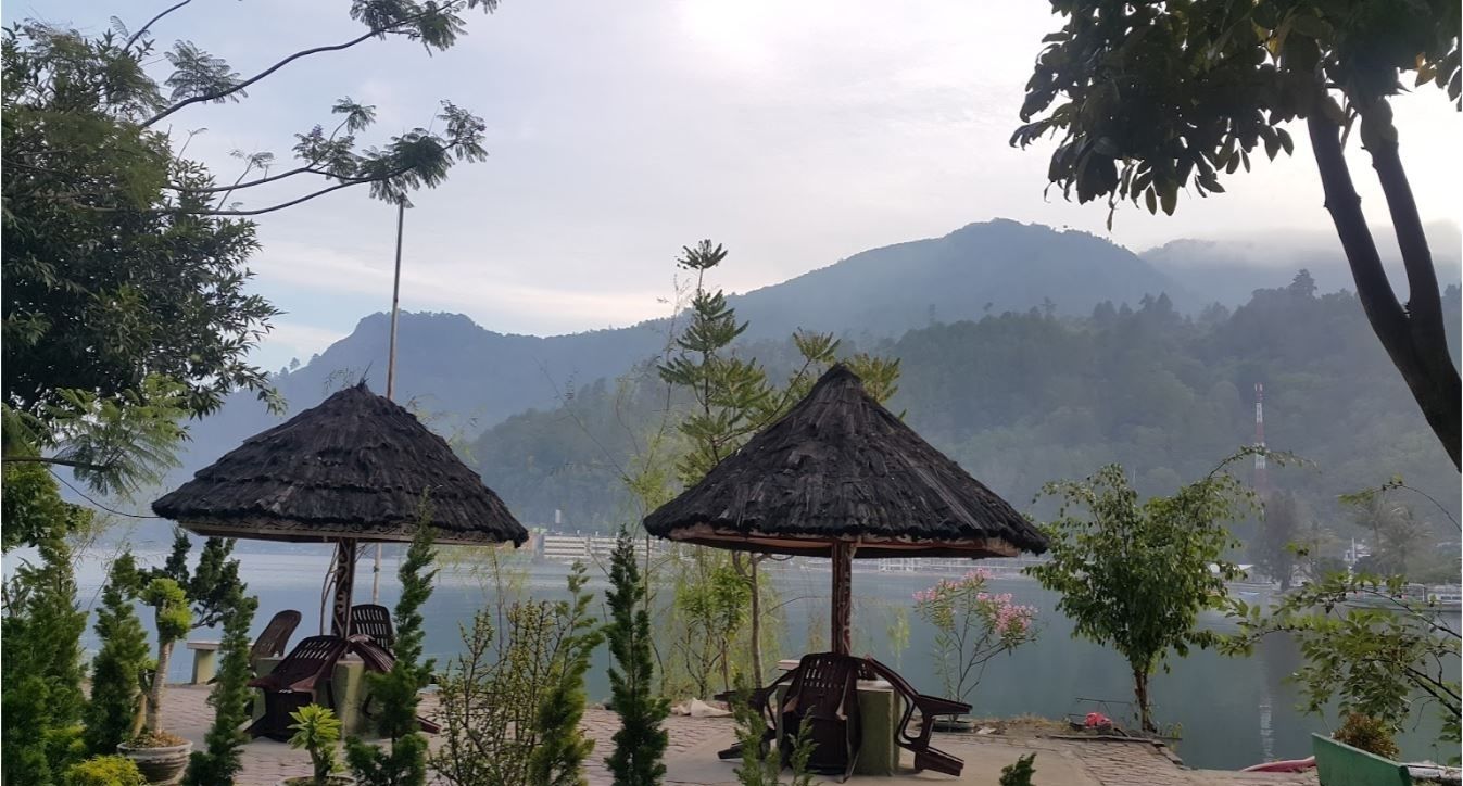 Um cenário tranquilo à beira de um lago, com gazebos de colmo e cadeiras, cercado por vegetação exuberante e montanhas ao fundo.