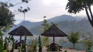 Um cenário tranquilo à beira de um lago, com gazebos de colmo e cadeiras, cercado por vegetação exuberante e montanhas ao fundo.