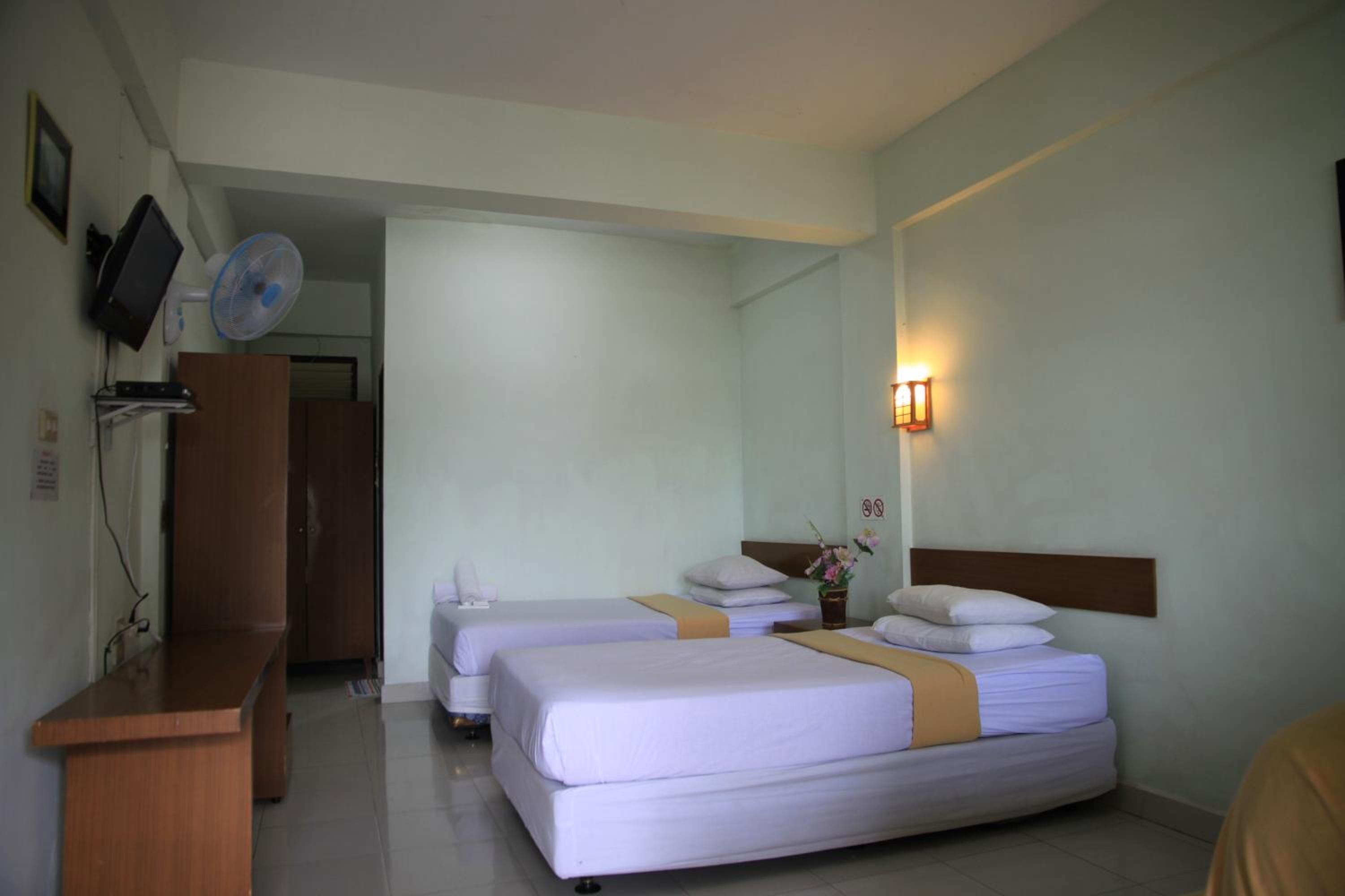 A hotel room with two beds, a desk, a TV, a wall-mounted fan, and a bedside lamp.