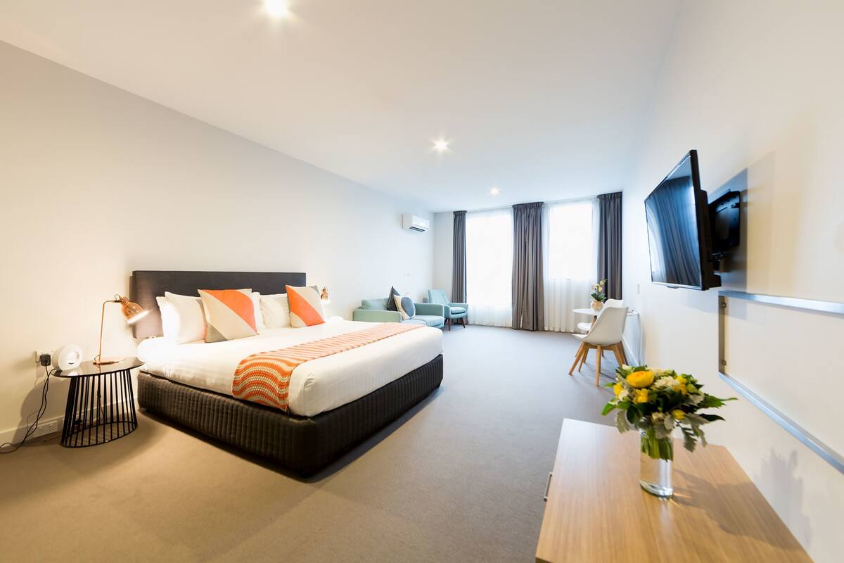 A modern hotel room with a large bed, a desk, a chair, a TV, and a vase of flowers.