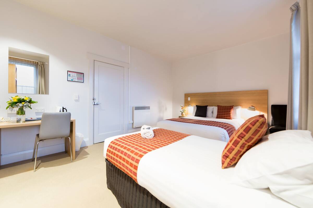 A hotel room with two beds, a desk, a chair, a small table with a vase of flowers, and a window with curtains.