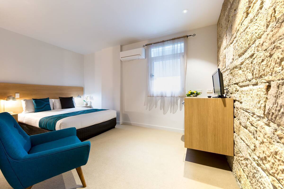 A modern hotel room with a bed, a blue armchair, a wooden desk, and a stone wall.