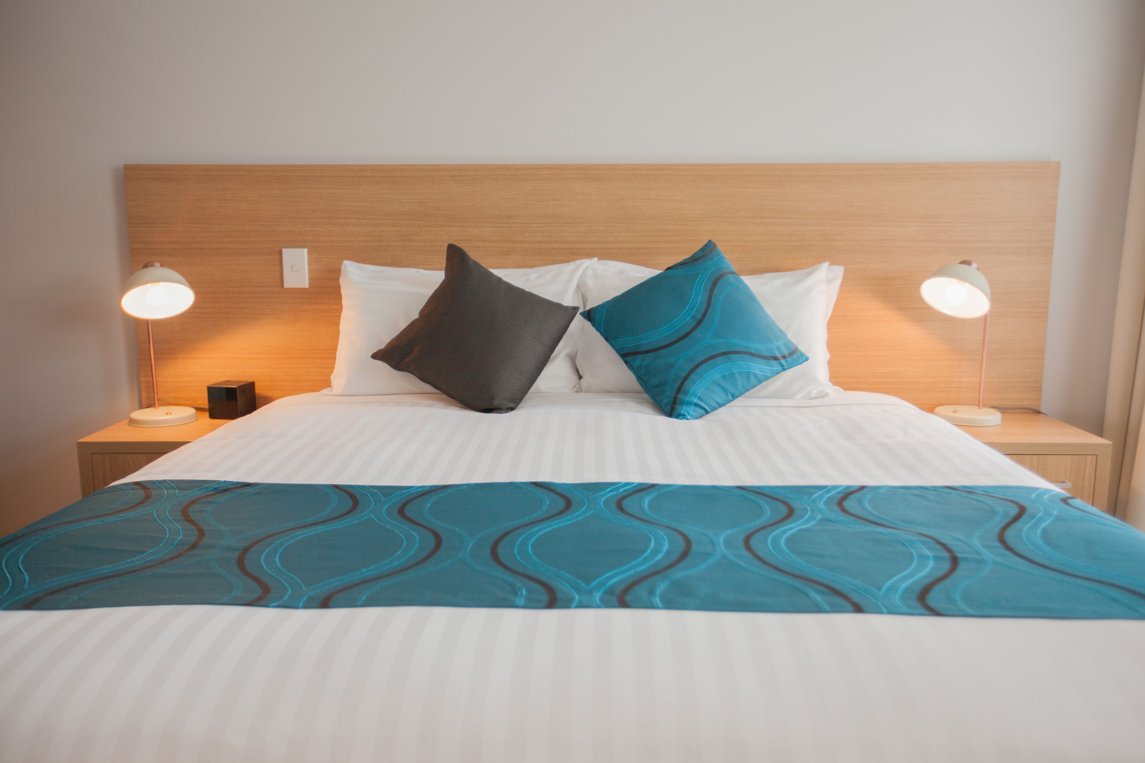 A neatly made bed with a white and teal bedspread, two bedside lamps, and a wooden headboard.
